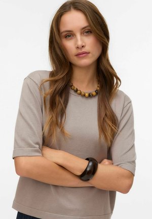 Femme aux longs cheveux bruns portant un haut à manches courtes beige, un collier de perles et un bracelet noir, les bras croisés, sur fond blanc.