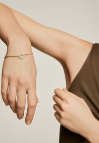 La pulsera dorada presenta un acento de piedra circular en un tono verde pálido. El diseño es delicado, con una cadena fina y una estética minimalista.