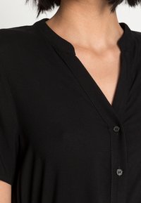 Schwarzes kurzärmliges Hemd mit V-Ausschnitt und drei Knöpfen. Glattes Material mit minimaler Textur. Dezentes Nähdetail entlang des Kragens.