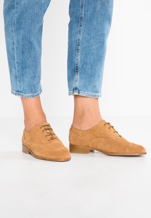 Brune oxfordsko i ruskind med lav hæl, som har snørebånd og en glat overdel med en syet toedækning. Båret med korte blå jeans.