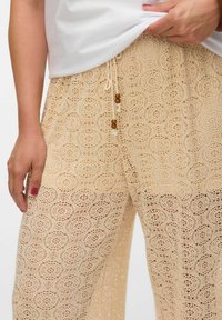 Beige heklet bukser med blomstermønster, snore med træperler og et løs, let design.
