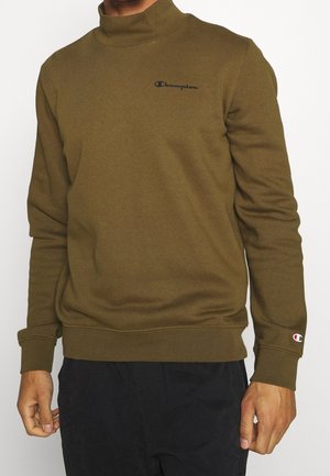 Mand iført olivengrøn Champion sweatshirt med høj krave og sorte bukser foran hvid baggrund.