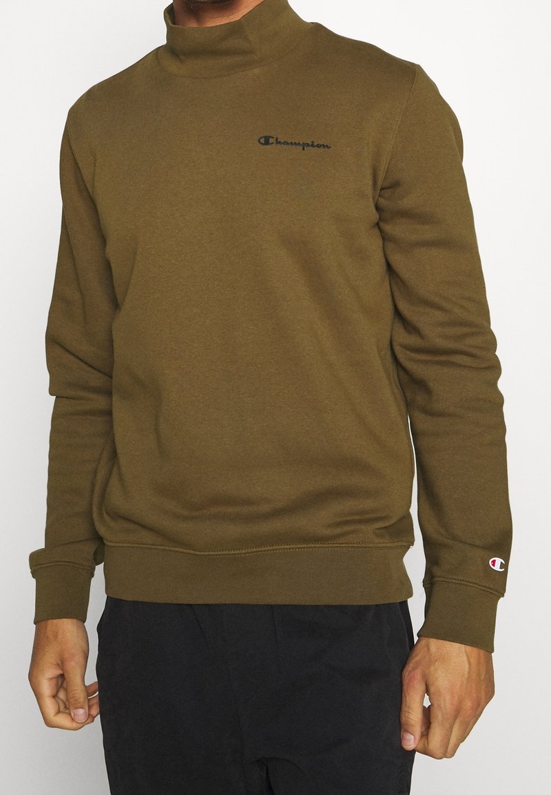 Homme portant un sweat-shirt Champion vert olive à col haut et un pantalon noir sur fond blanc.