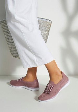 Helle rosa, Netz-Sneaker mit Schnürung und weißen Sohlen, kombiniert mit weiten weißen Hosen und einer geflochtenen Tasche, vor einem neutralen Hintergrund.