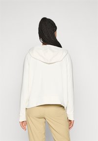 Person med langt fletthår som har på seg en hvit hettegenser og beige bukser, stående med ryggen mot en ensfarget lys bakgrunn.
