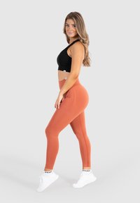 Zwarte sportbeha en roestkleurige leggings van textuurstof, met een hoge tailleband en witte sneakers. Model staat zijwaarts.