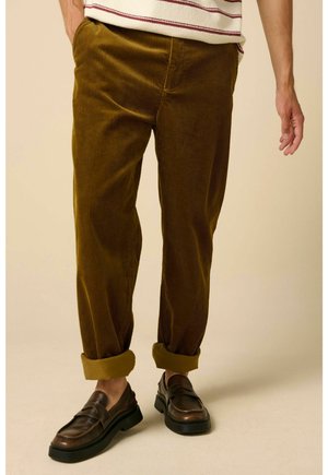 Man wearing rolled-up brown corduroy pants and dark brown leather loafers standing on a beige surface.