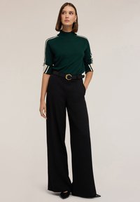 Pulover verde cu guler înalt, având dungi albe pe mâneci, asortat cu pantalonii negri cu talie înaltă și linii largi, împreună cu o curea neagră cu cataramă aurie.