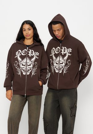 Deux personnes portent des sweats à capuche zippés marron assortis avec un grand visage de démon blanc et le texte ""NOPE"", associés à un pantalon sombre usé.