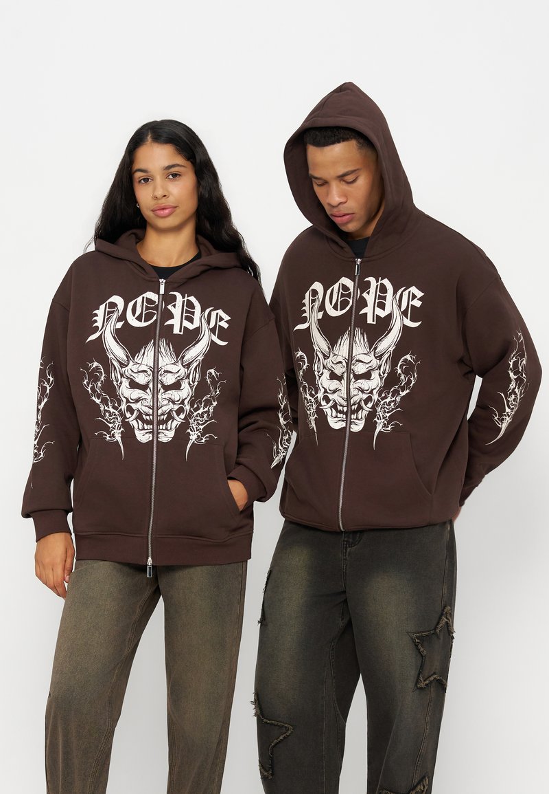 Deux personnes portent des sweats à capuche zippés marron assortis avec un grand visage de démon blanc et le texte ""NOPE"", associés à un pantalon sombre usé.