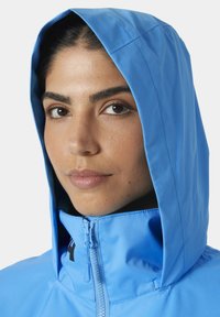 Blauwe hooded jacket met een hoge kraag, ritssluiting aan de voorkant en een gladde textuur. Opvallende details zijn onder meer een logo op de borst en gestikte randen.