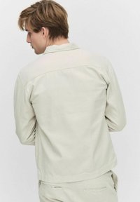 Beige katoenen jack met een rechte snit, puntkraag en details op de rug. Heeft lange mouwen en een minimalistisch ontwerp.