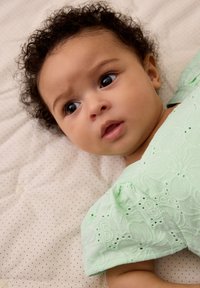 Mintgroene jurk met bloemenpatroon, korte pofmouwen, rustend op een stippendekentje. Baby met krullend donker haar en grote ogen.
