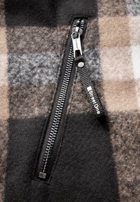 Giacca in pile con zip, caratterizzata da un motivo a quadri nero, grigio e marrone, con una zip e una texture del tessuto a contrasto. Etichetta del marchio visibile.