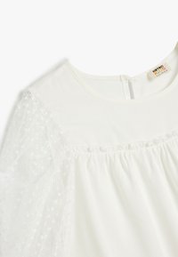 Weiße Bluse mit puffartigen transparenten Ärmeln im Punktmuster, runder Halsauschnitt und einer Knopfverschluss hinten. Weicher, leichter Stoff.