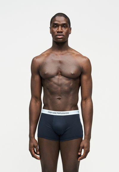 Atletyczny mężczyzna o ciemnej karnacji stojący przodem, ubrany w czarne bokserki Emporio Armani na jednolitym białym tle.