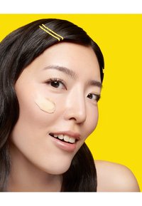 Produit de fond de teint appliqué sur la joue d'une personne aux cheveux raides et foncés, portant des pinces à cheveux jaunes, sur un fond jaune brillant.