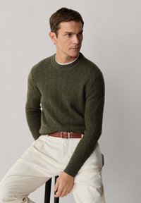Maglione a coste verde oliva con scollo tondo, indossato sopra una camicia bianca, abbinato a pantaloni crema e una cintura in pelle marrone.