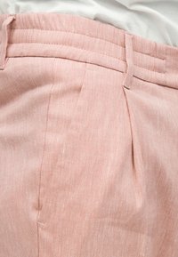 Gros plan sur un pantalon en lin rose clair avec passants de ceinture, plis et un haut blanc partiellement visible rentré à l'intérieur.
