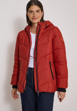 Femme portant une veste matelassée rouge à capuche par-dessus une chemise blanche et un jean foncé, debout devant un fond clair uni.