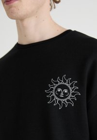 Felpa nera con un design solare ricamato bianco sul lato sinistro del petto, che mostra un volto e raggi che irradiano. Materiale in cotone, stile a girocollo.