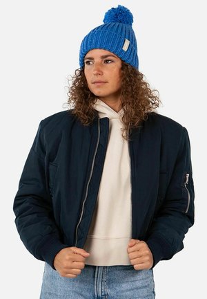 Marineblaue Bomberjacke mit gerippten Bündchen, Reißverschlusstaschen und einem cremefarbenen Hoodie darunter, kombiniert mit einer gestrickten blauen Mütze mit Pompon.