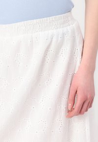 Jupe blanche avec un motif floral texturé, dotée d'une taille élastique froncée. Le tissu présente des broderies complexes et des découpes.
