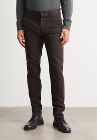Brown cotton trousers with a slim fit, featuring a button closure and side pockets, paired with black lace-up shoes on a gray surface.