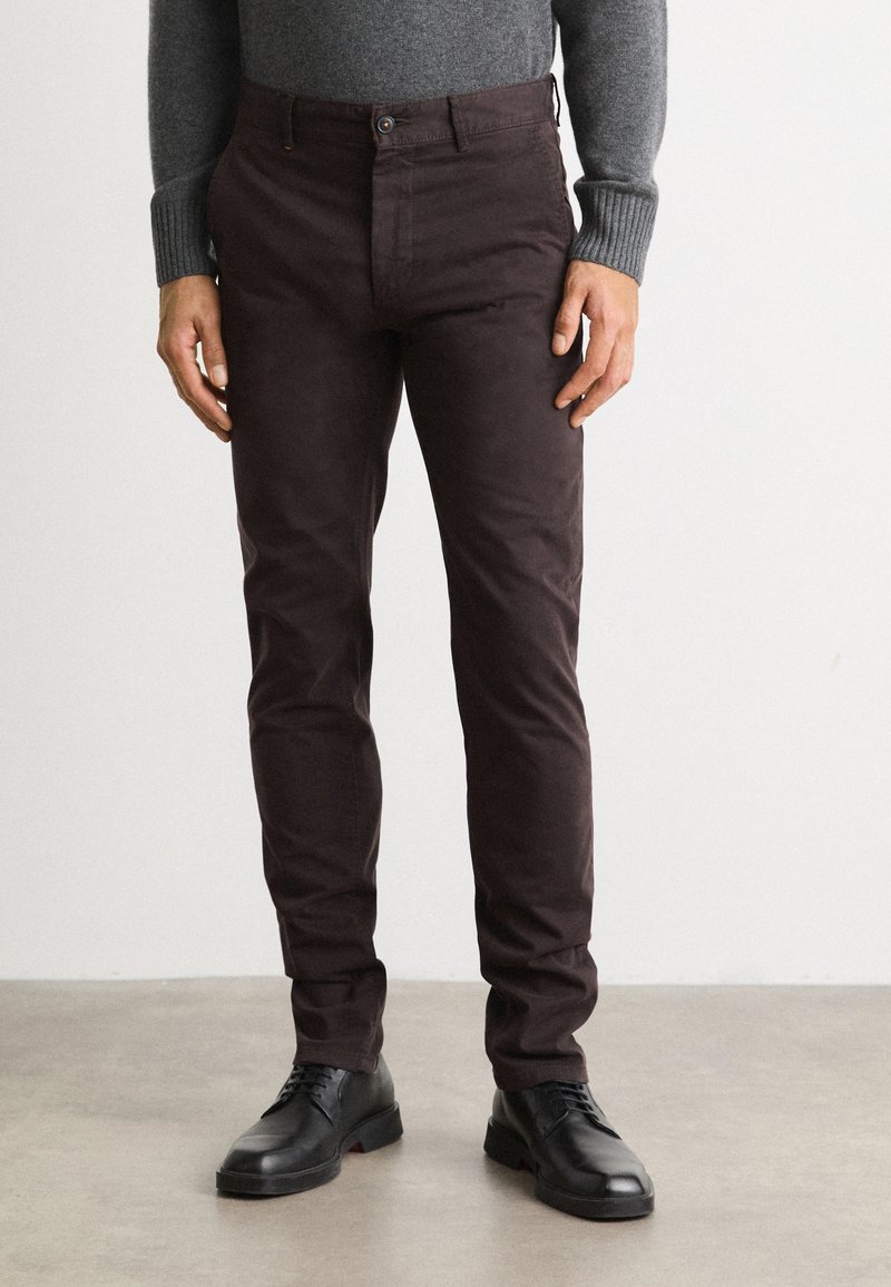 Brown cotton trousers with a slim fit, featuring a button closure and side pockets, paired with black lace-up shoes on a gray surface.