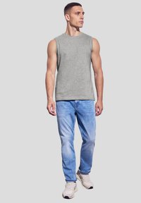 Graues ärmelloses Shirt, hellblaue Jeans und beiges Sneakers. Mann steht vor einem neutralen Hintergrund und präsentiert die Details seines lässigen Outfits.
