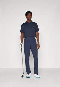 Μπλε σκούρο πόλο μπλουζάκι με κοντά μανίκια και κίτρινο λογότυπο, σε συνδυασμό με μπλε σκούρο παντελόνι. Φορεμένο με λευκά παπούτσια γκολφ και κρατώντας ένα γκολφ ρόπαλο.