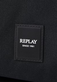 Schwarzes Stoffetikett mit strukturiertem Oberflächen, das den Markennamen "REPLAY" und "SINCE 1981" in weißem Text auf schwarzem Hintergrund zeigt.