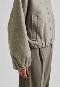 Lys grå, teksturert fleecejakke med glidelås foran, elastiske mansjetter og en sidelomme, kombinert med olivengrønne bukser.