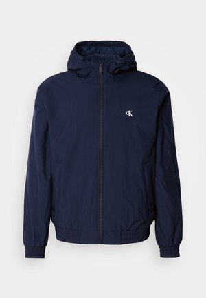 Gaiši tumši zils viegls džakets ar pilnu rāvējslēdzēju, kapuci, elastīgām aprocēm un jostasvietu. Kreisajā krūtīs mazs balts logotips.