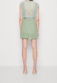 Blouse à dos en dentelle de couleur bleu clair avec des manches volantées, associée à une jupe en olive vert doux, plissée jusqu'au genou, et des chaussures à talons neutres.