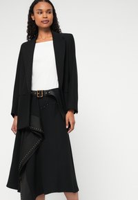 Melns žakets ar platiem klāja ieliktņiem, apvienots ar jostu ar piedurkņu apakšmalu un studu akcentiem. Zem tā redzams balts tops.