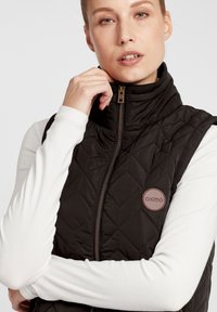 Svart quiltad väst med hög krage, prydd med guldzipförslutning och en rund brun logotyp. Modellen bär en vit långärmad tröja under.