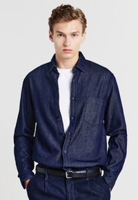 Camicia di jeans navy con bottoni, maniche lunghe, taschino sul petto e una texture discreta, indossata sopra una t-shirt bianca e una cintura nera.