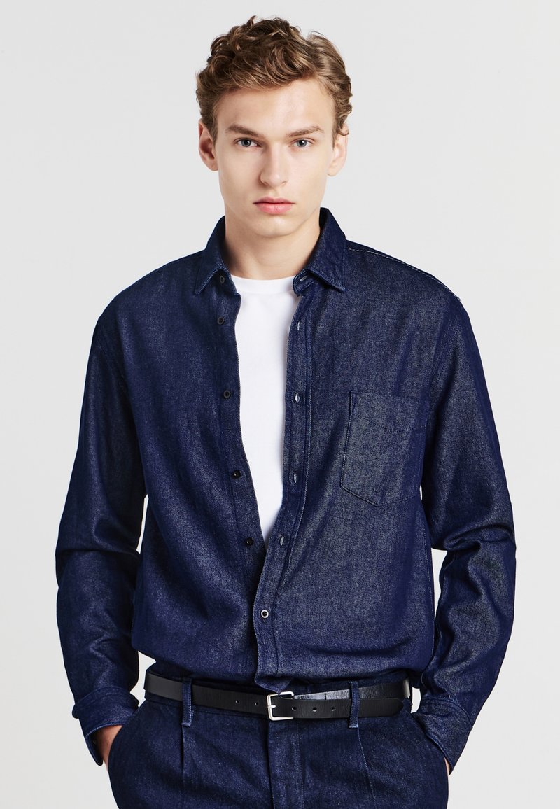 Camicia di jeans navy con bottoni, maniche lunghe, taschino sul petto e una texture discreta, indossata sopra una t-shirt bianca e una cintura nera.