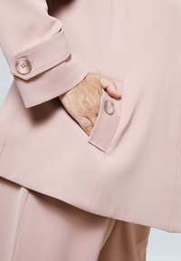 Light pink blazer sleeve with two-button cuff detail. Pocket with a visible hand inside. Smooth fabric texture. Tailored fit.