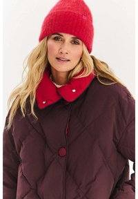 Bordowa pikowana kurtka z czerwonym kołnierzem i guzikami zapinającymi; wykonana z miękkiego, teksturowanego materiału. W zestawie czerwona dziana czapka.