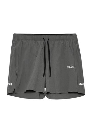 Mand stående iført en sort T-shirt med lille tekst på brystet, grå teksturerede shorts, hvide sokker og grå sneakers på hvid baggrund.