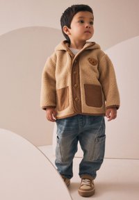Veste en polaire beige avec des accents marron, fermeture à boutons, deux poches avant. Assortie à un pantalon en denim bleu et des baskets beiges. Pose centrale.
