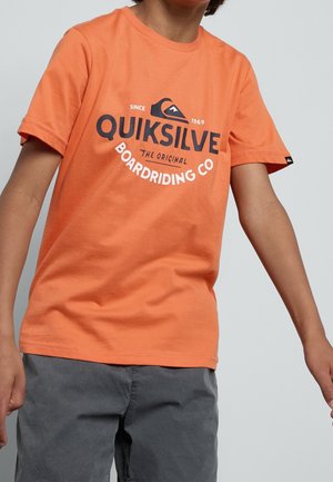 Dziecko ubrane w pomarańczowy t-shirt Quiksilver z logo i napisem, połączony z szarymi szortami, stojące na neutralnym tle.