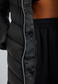 Zwarte pufferjack met een geluiddichte ontwerp, glanzende afwerking en een volle lengte rits met een contrasterende logo-detail langs de sluiting.