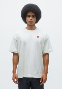 Light grey oversized t-shirt with short sleeves, featuring a small red logo on the left chest. Smooth cotton material, straight cut, casual fit.
