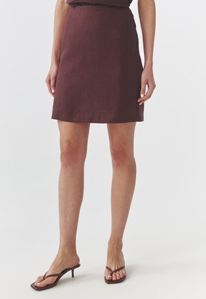 Jupe en lin bordeaux avec une coupe droite et des détails de coutures minimalistes, associée à des tongs à talons marron. Texture lisse et longueur au genou.