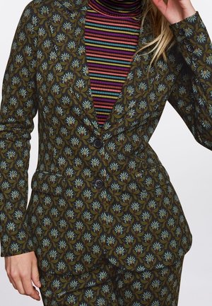 Donna che indossa un completo verde a motivi floreali con due bottoni sopra un dolcevita a righe multicolore.