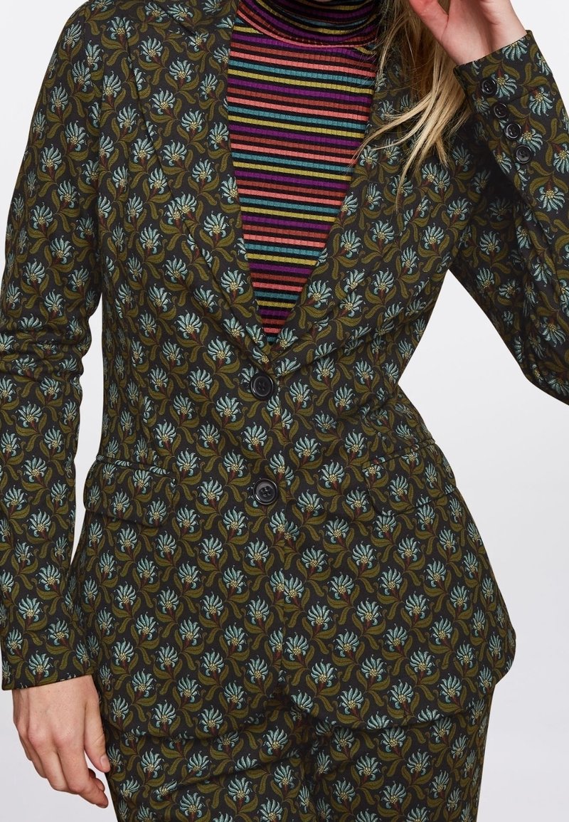 Femme portant un costume vert à motif floral avec deux boutons sur un pull à col roulé multicolore rayé.