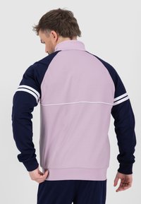Giacca sportiva viola chiaro e blu navy con colletto zip, maniche raglan e dettagli a strisce bianche. Tessuto liscio e design aderente.